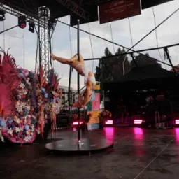 Akrobatka w białym stroju wykonuje figurę na rurze, na scenie z dekoracjami kwiatowymi i oświetleniem scenicznym. Widoczny zespół muzyczny w tle. Nietypowe ujęcie.