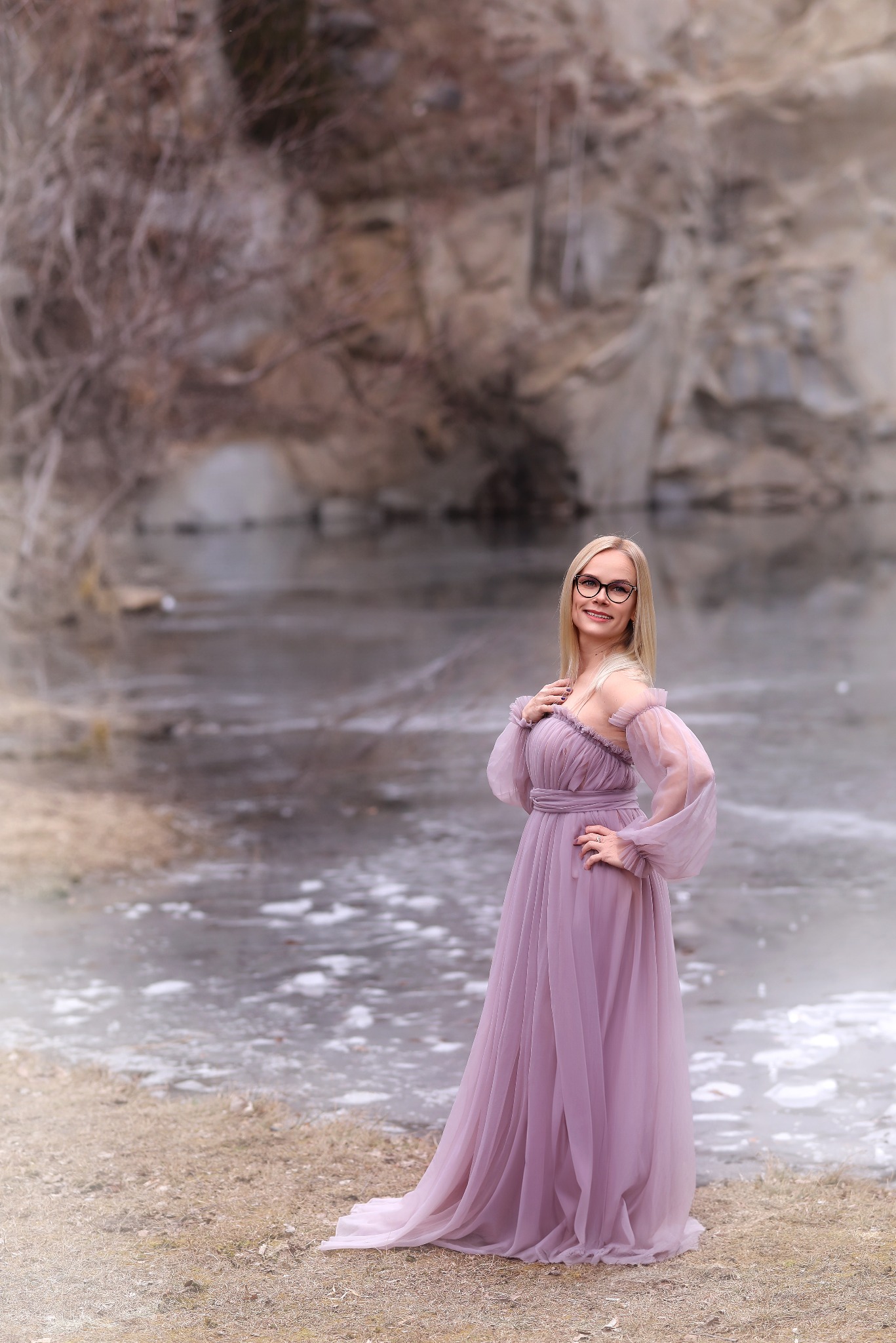 Kobieta w długiej, liliowej sukni pozuje na tle zamarzniętego jeziora i skalistego krajobrazu. Uśmiechnięta blondynka w okularach, z ręką na biodrze, prezentuje elegancką kreację.