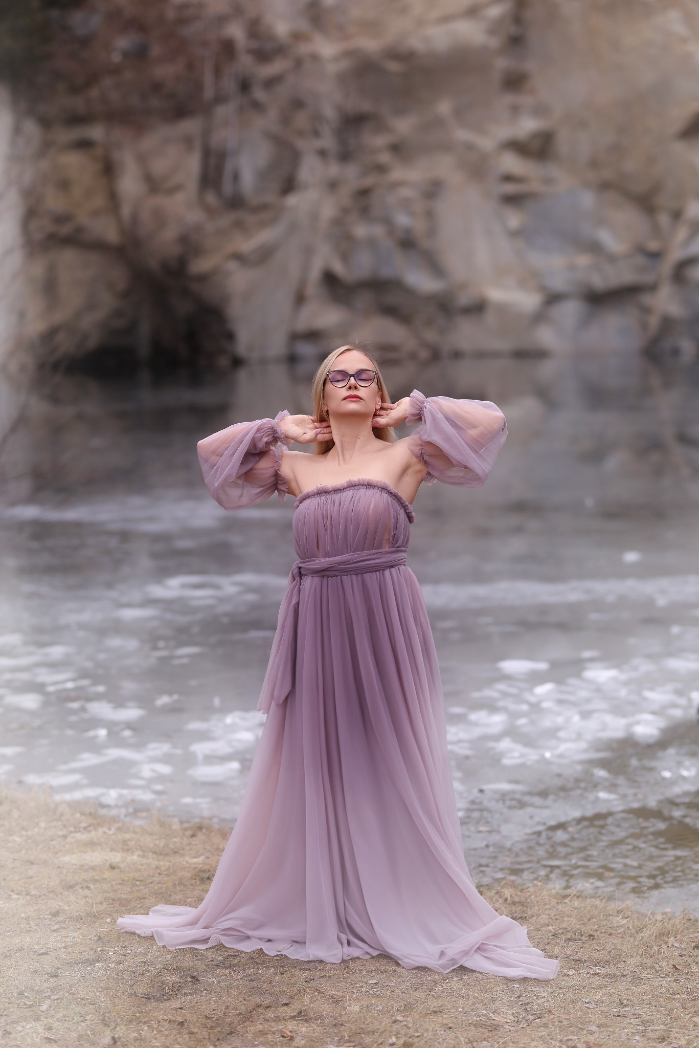 Kobieta w długiej, zwiewnej sukni w odcieniach fioletu i okularach, pozuje na tle skalistego krajobrazu z zamarzniętym jeziorem, ręce uniesione za głową.