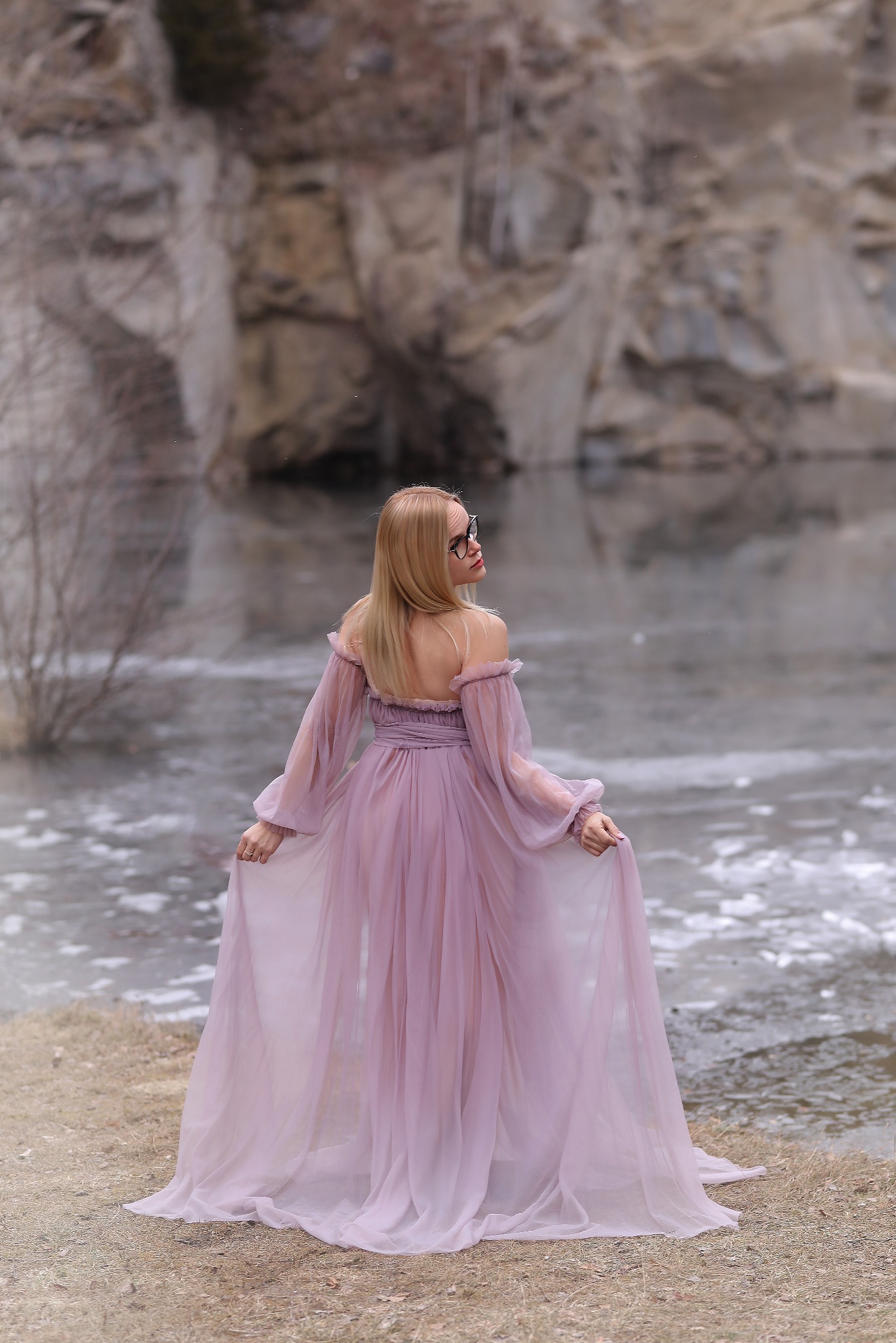 Kobieta w zwiewnej, liliowej sukni, odwrócona tyłem, na tle skalnego krajobrazu i zamarzniętego jeziora. Romantyczna sceneria, delikatne światło, sesja w plenerze.