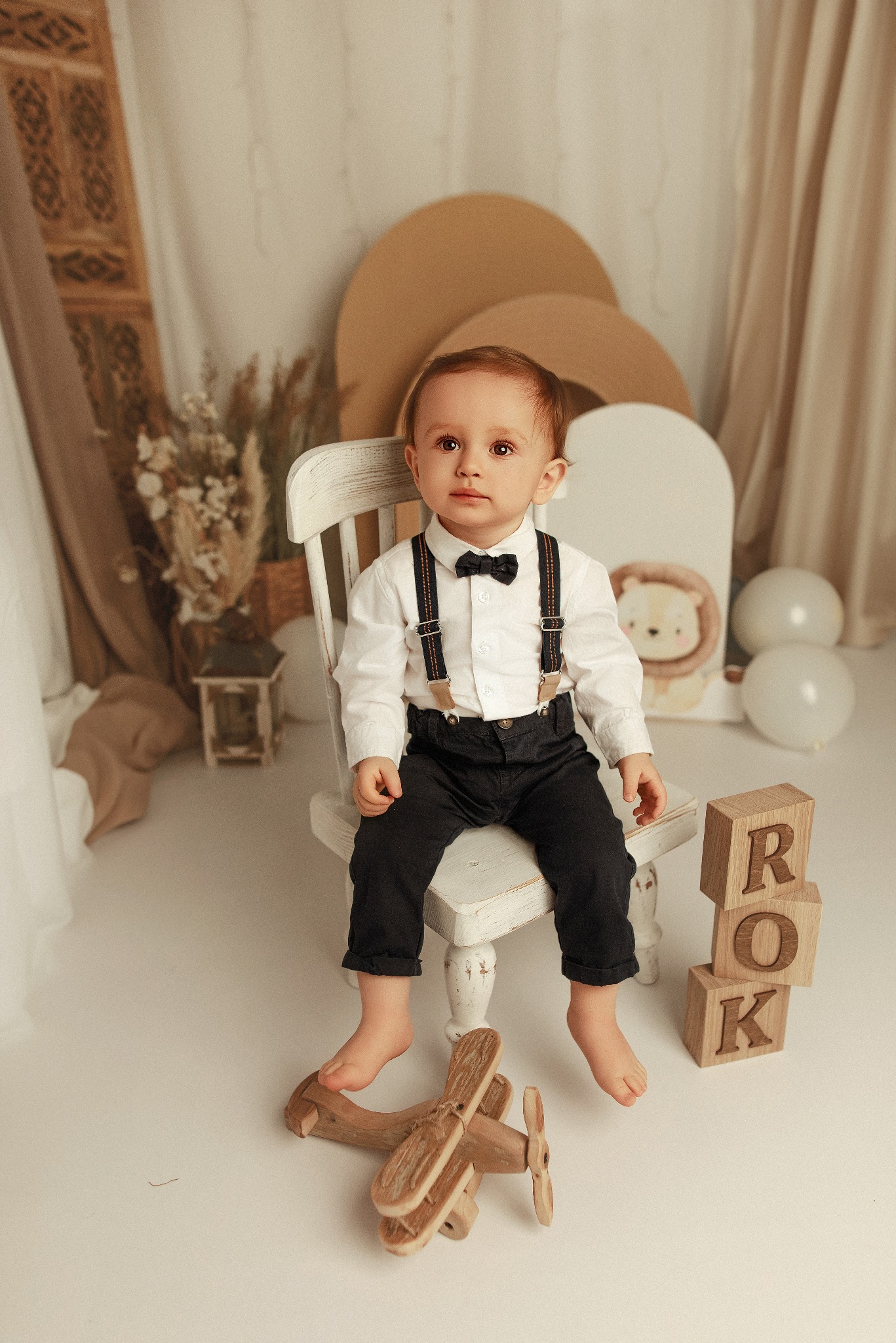 Uroczy chłopiec w muszce i szelkach siedzi na białym krześle, obok drewniany samolot i klocki z napisem ROK, w tle dekoracje w neutralnych barwach.