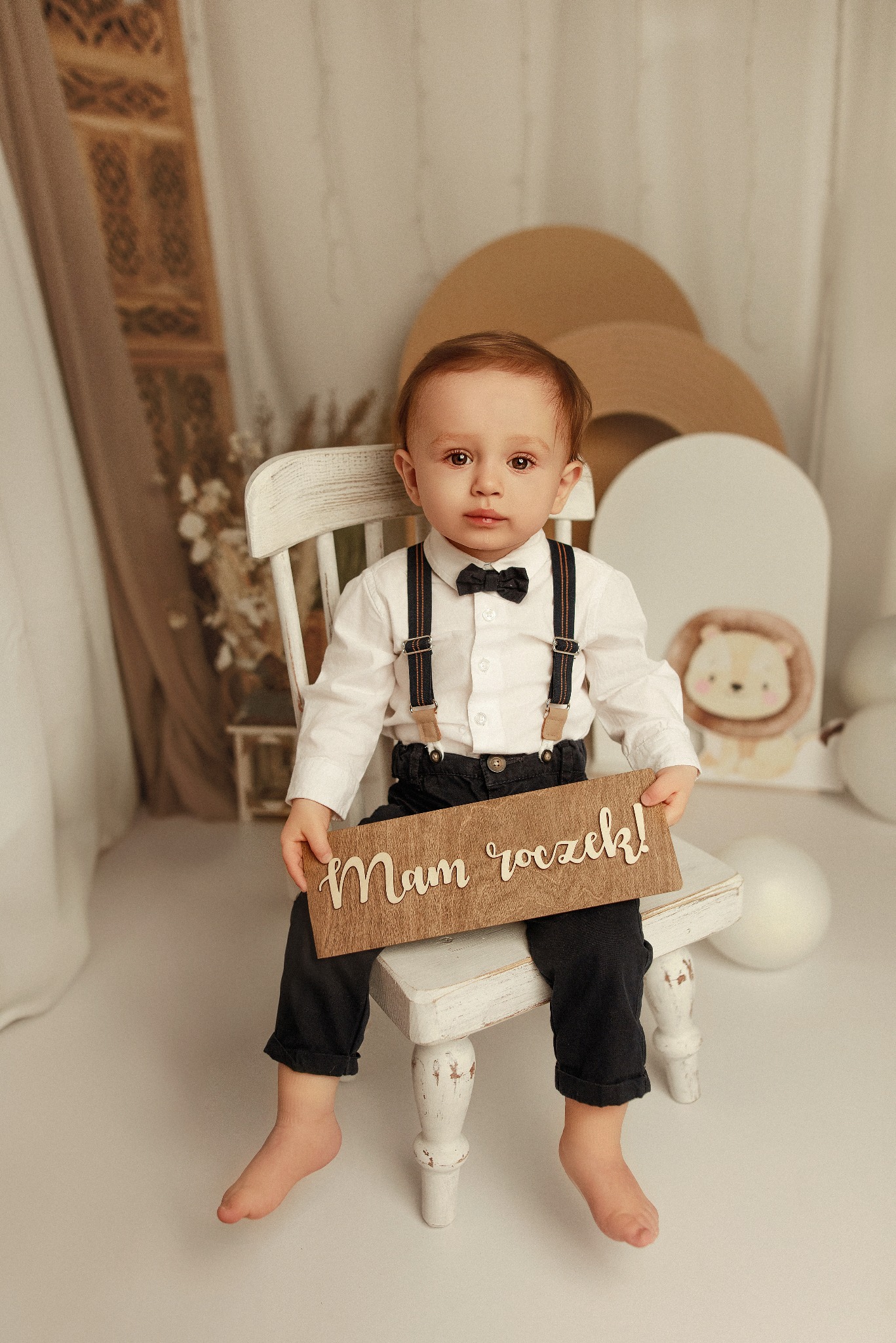 Roczne dziecko w eleganckim stroju siedzi na krześle, trzymając drewniany napis 'Mam roczek!', w tle dekoracje urodzinowe i portret lwa.