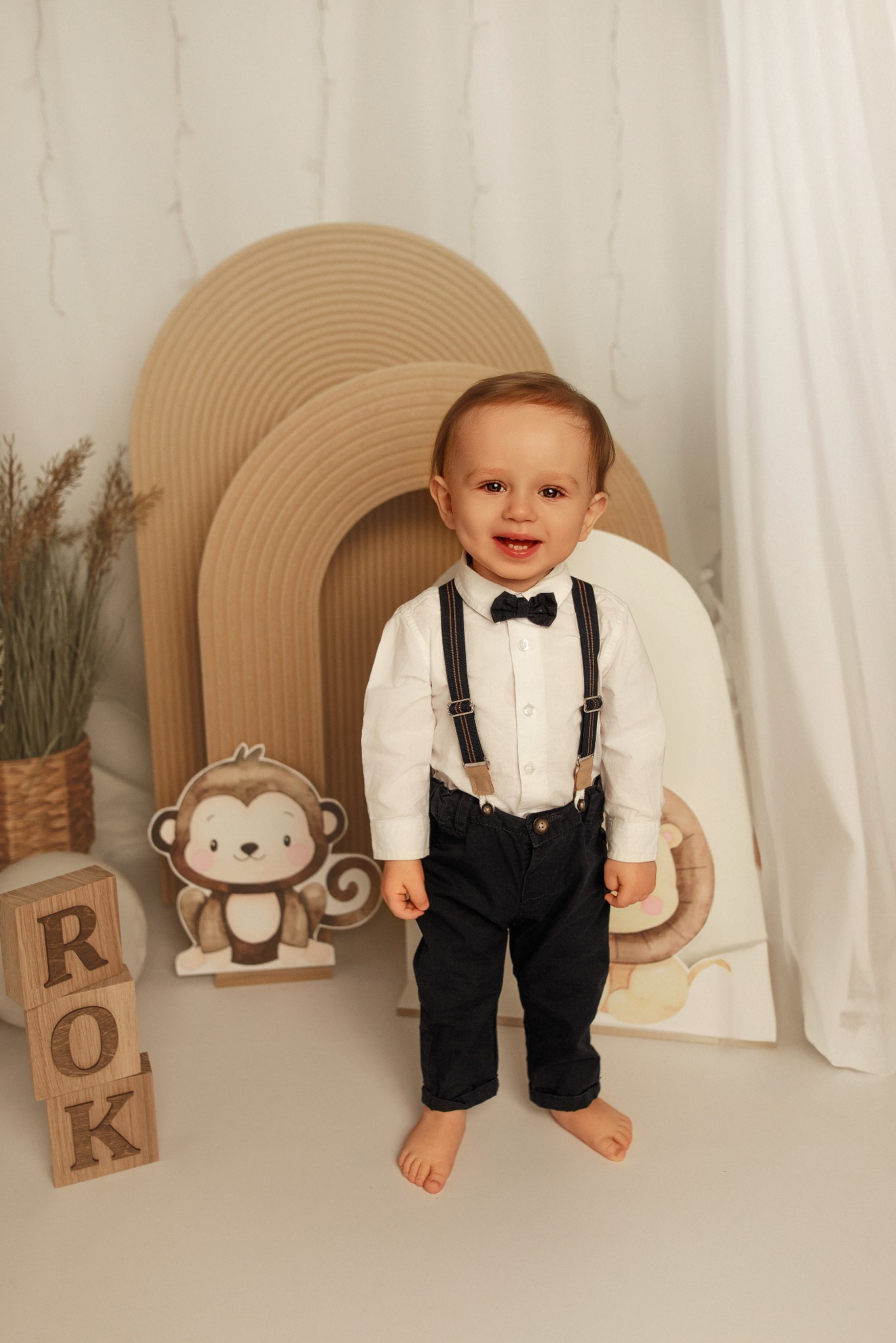 Uśmiechnięty roczny chłopiec w koszuli, muszce i szelkach stoi na bosaka na tle dekoracji urodzinowych z motywami zwierzęcymi i drewnianymi klockami z napisem 'ROK'.
