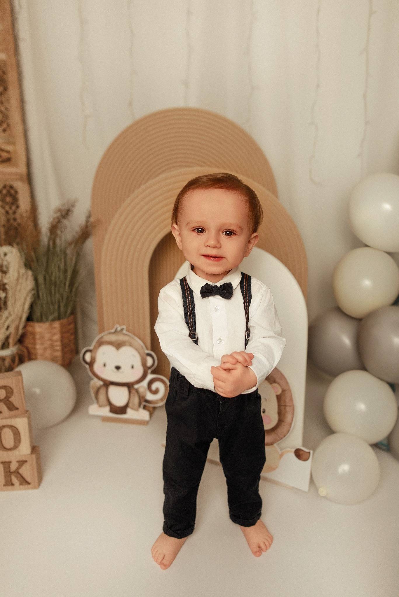 Radosny roczek w studio: Uroczy chłopiec w muszce i szelkach na tle dekoracji z motywem zwierzątek i balonów, świętuje swoje pierwsze urodziny.