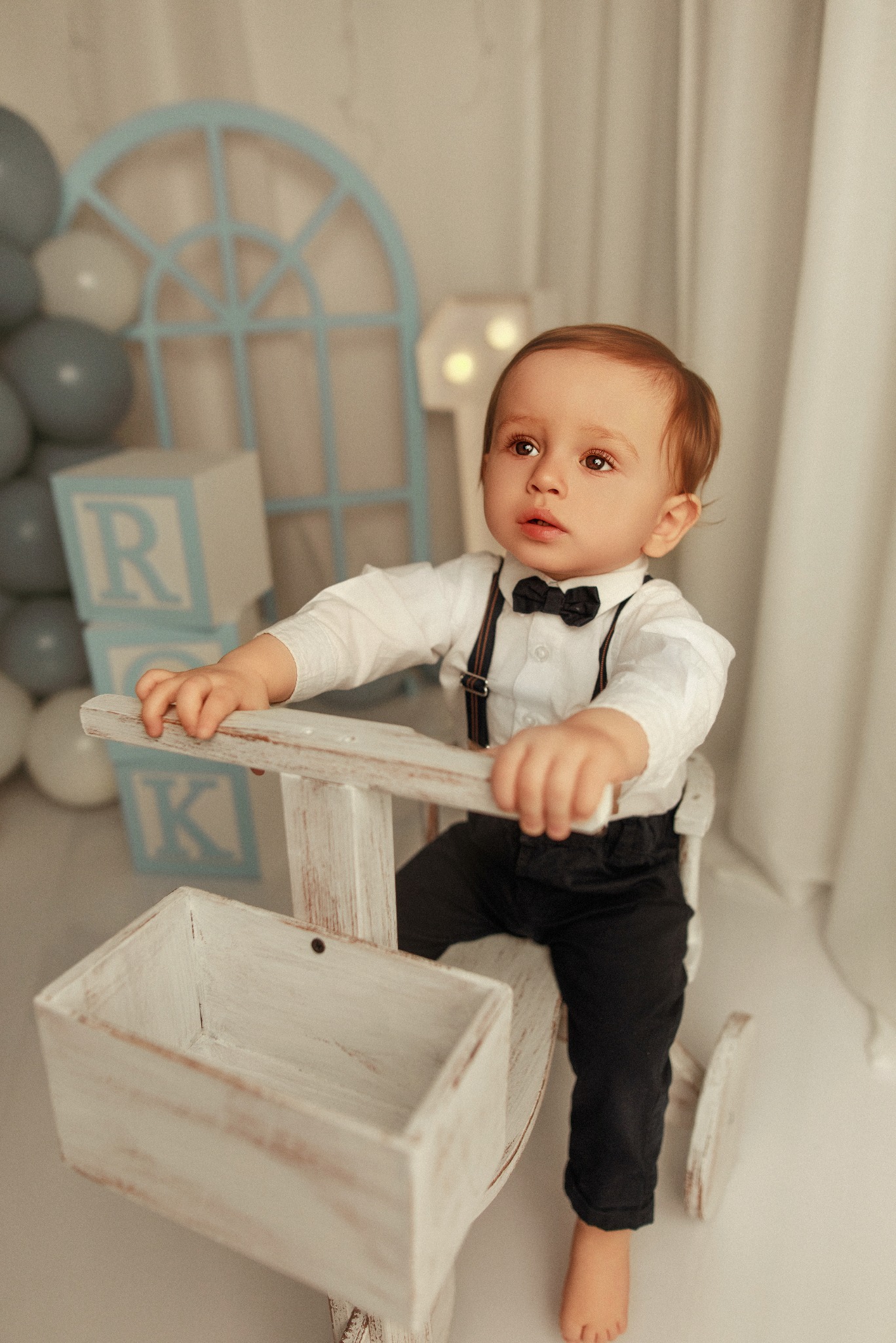 Uroczy chłopiec w muszce i szelkach siedzi na drewnianym koniku na biegunach w pastelowej scenografii z balonami i klockami z literami.