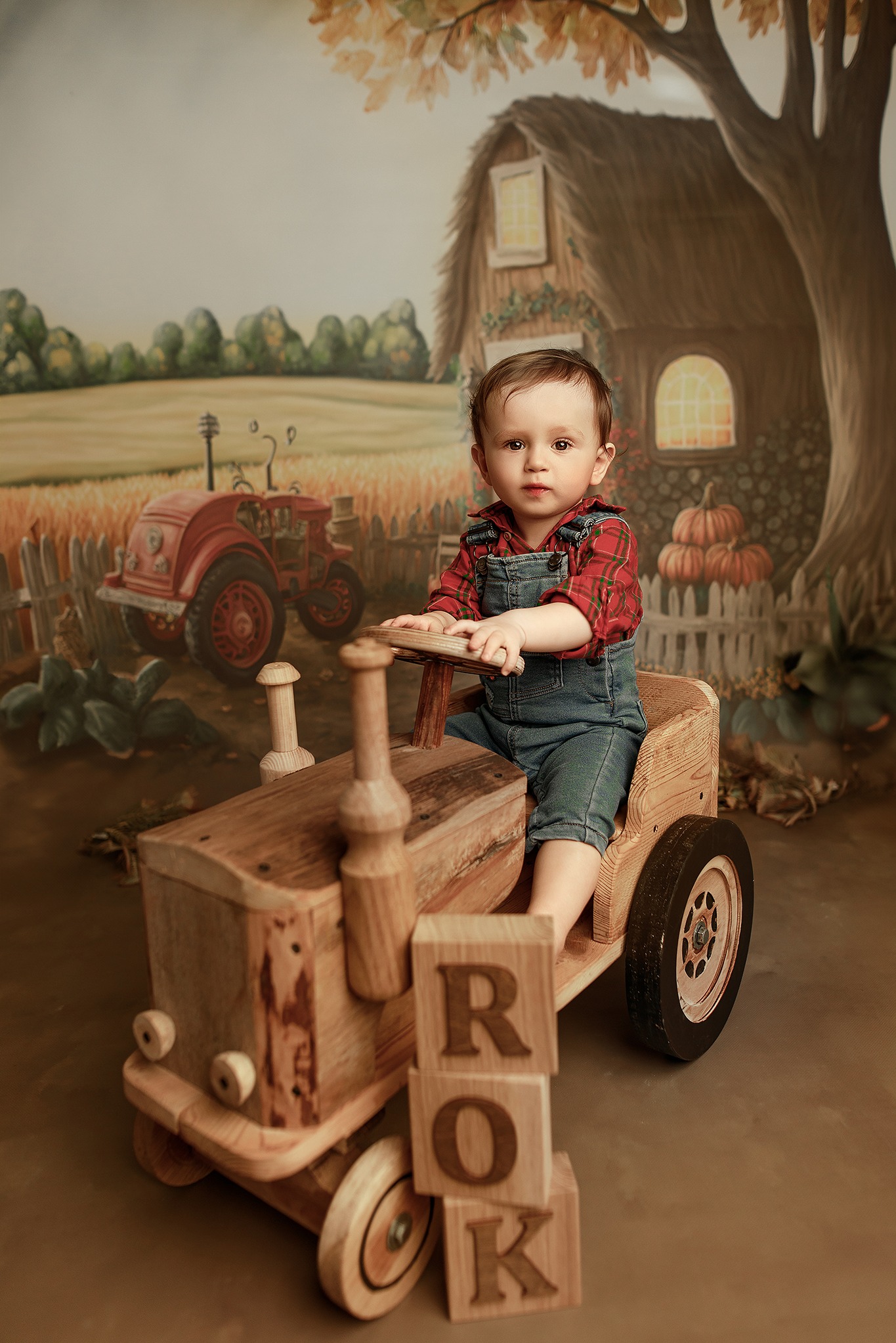 Uroczy chłopiec w dżinsowym kombinezonie i kraciastej koszuli siedzi w drewnianym traktorze na tle malowanej scenerii wiejskiej.