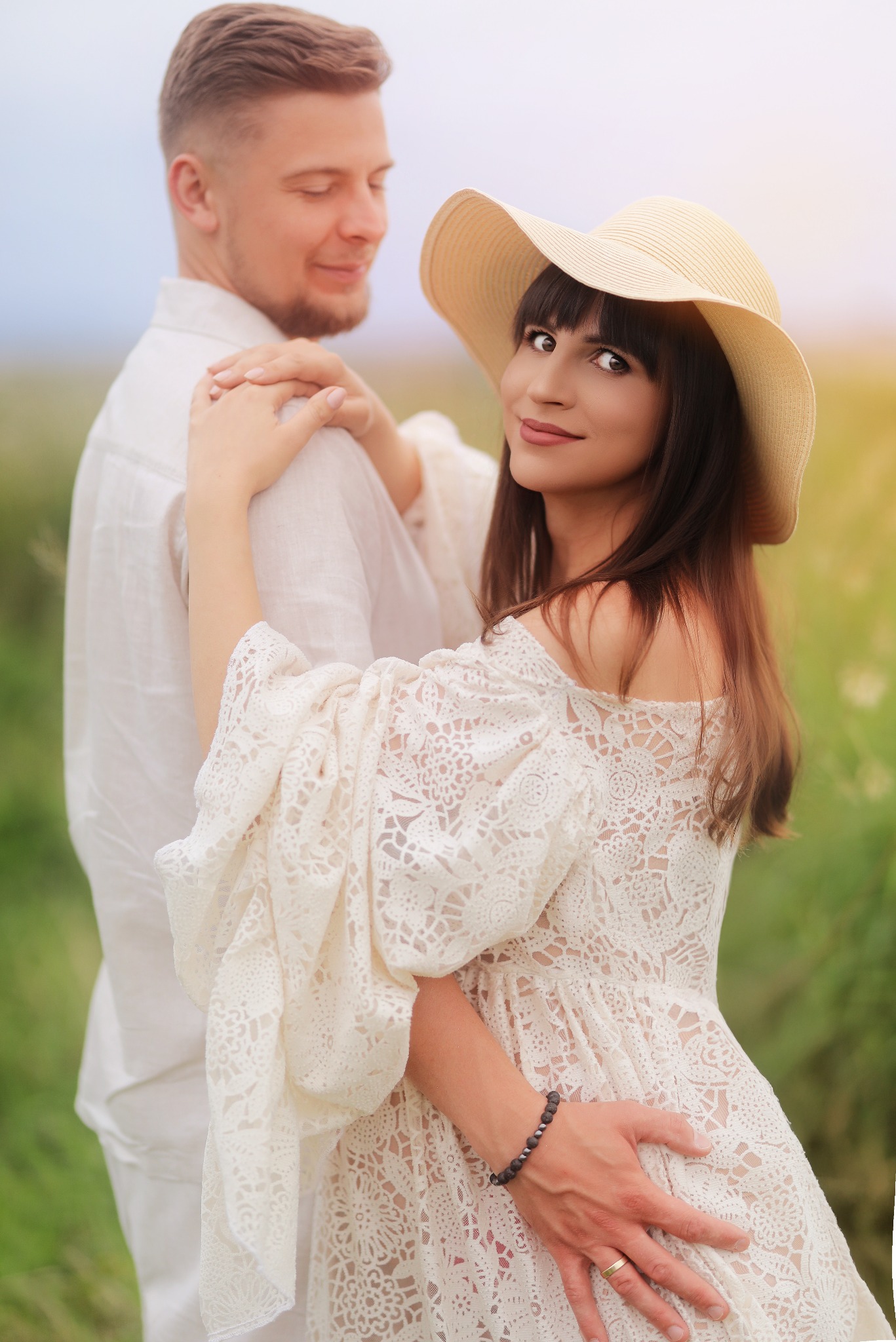 Uśmiechnięta kobieta w kapeluszu i koronkowej sukience, obejmowana przez mężczyznę w białej koszuli na tle łąki w słoneczny dzień. Romantyczna sesja narzeczeńska.