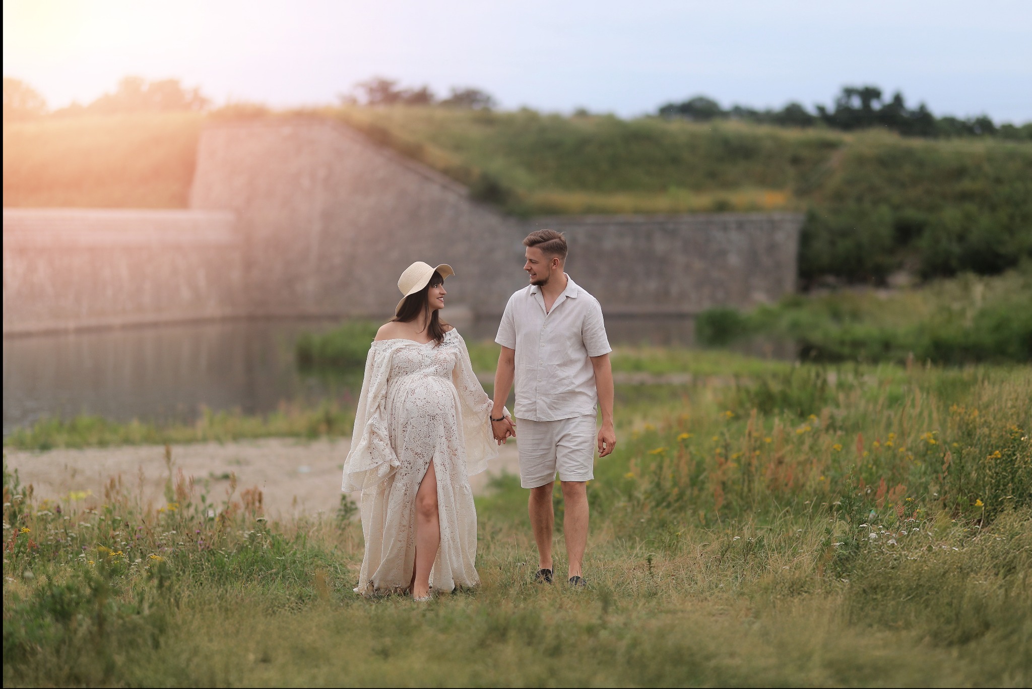 Sesja ciążowa w Otmuchowie: kobieta w koronkowej sukni i kapeluszu trzyma za rękę mężczyznę na tle muru i zbiornika wodnego w plenerze.