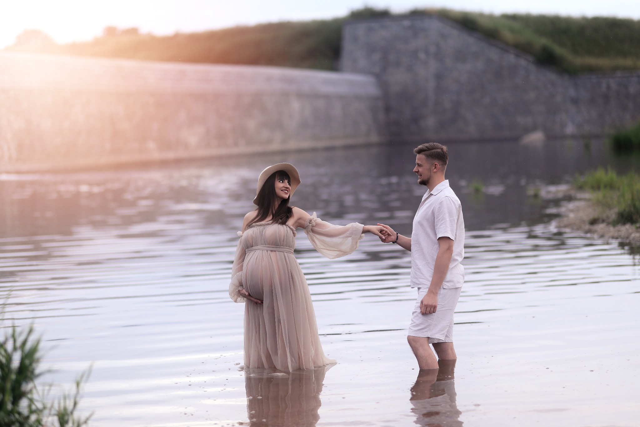 Uśmiechnięta kobieta w ciąży w zwiewnej sukni i kapeluszu trzyma za rękę mężczyznę, stojąc w wodzie na tle kamiennego muru i zieleni. Romantyczna sesja w plenerze.