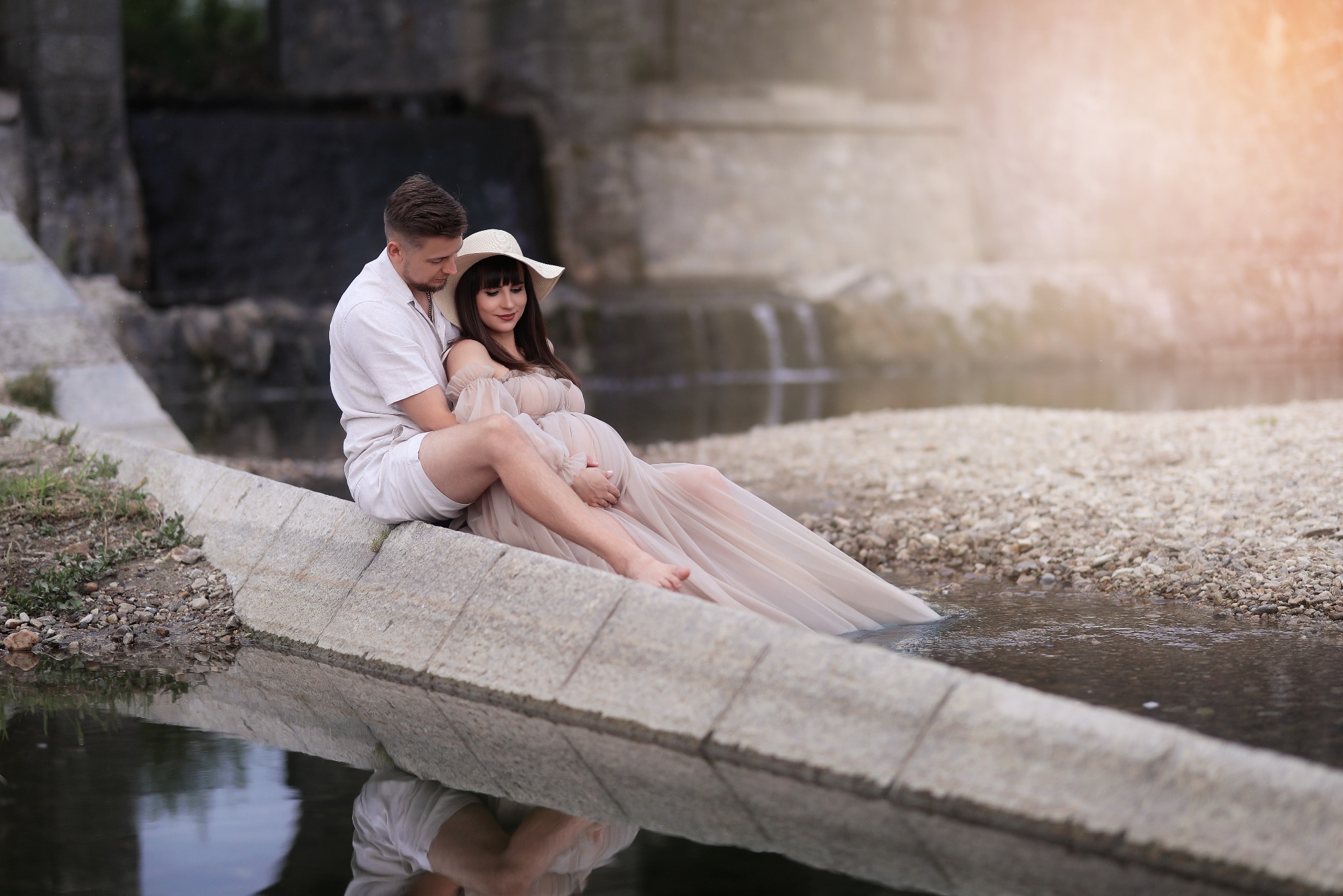 Uśmiechnięta, ciężarna kobieta w kapeluszu i zwiewnej sukni, siedzi z partnerem na betonowym brzegu płytkiej rzeki, w tle kamienny most i delikatne światło.