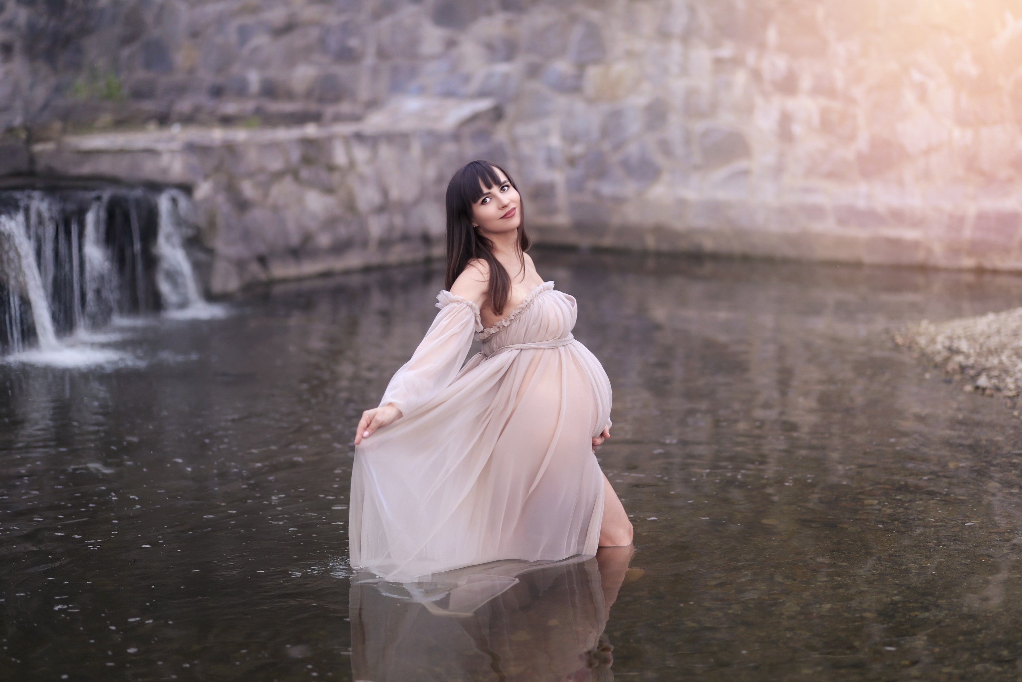 Kobieta w ciąży w zwiewnej sukni pozuje w płytkiej rzece z wodospadem i kamiennym brzegiem w tle, delikatne światło podkreśla naturalność scenerii.