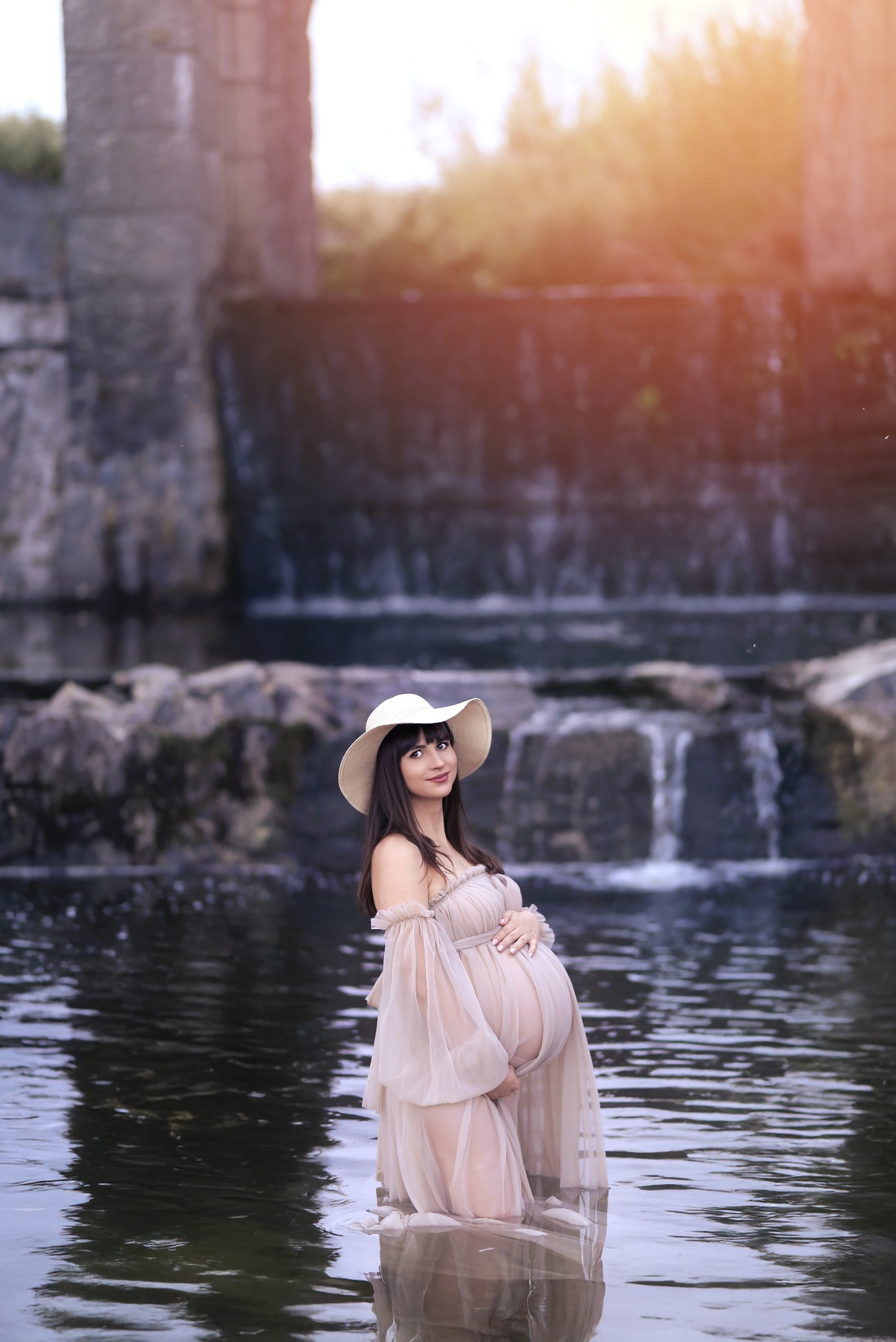 Kobieta w ciąży, w zwiewnej sukience i kapeluszu, pozuje w wodzie na tle wodospadu, w słoneczny dzień. Ujęcie pełne spokoju i oczekiwania.