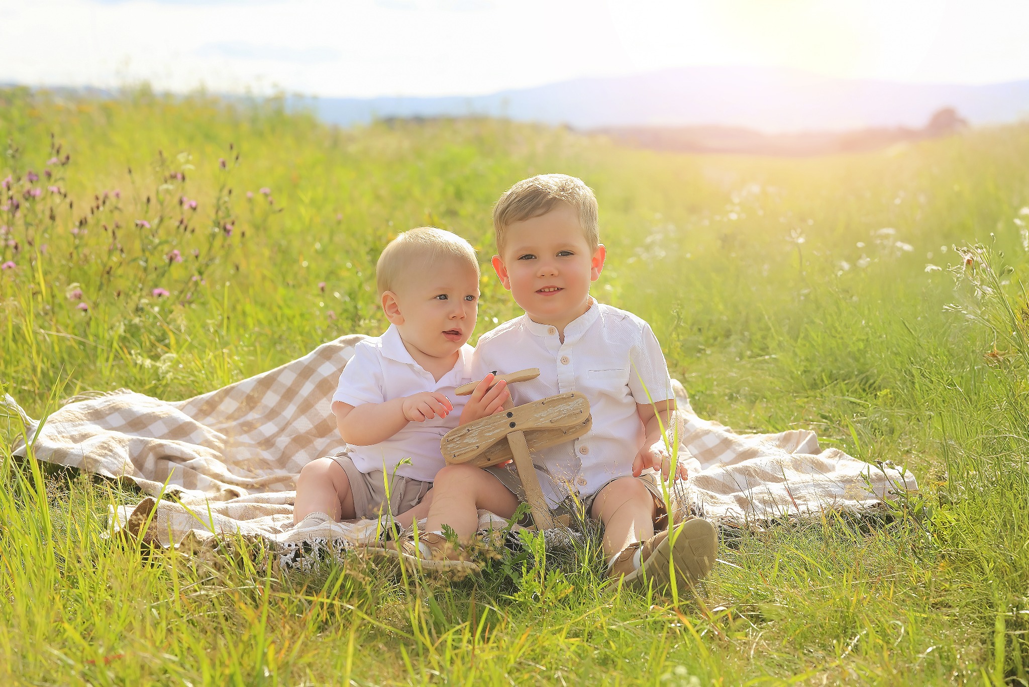 Dwóch chłopców w białych koszulach siedzi na kocu w trawie, trzymając drewniany model samolotu. W tle łąka i delikatne światło słoneczne.