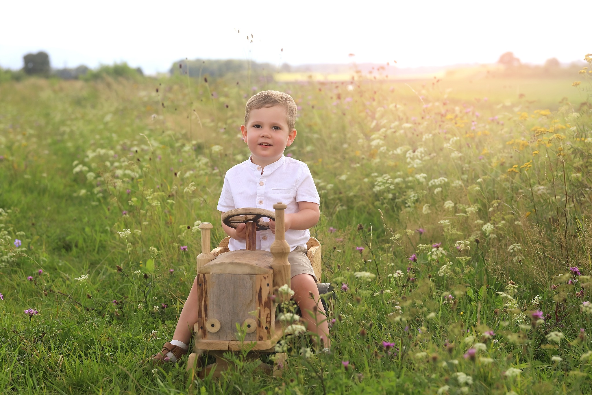 Chłopiec w białej koszuli siedzi na drewnianym traktorze na łące pełnej kwiatów w ciepłym świetle. Uśmiechnięta twarz dziecka skierowana w stronę obiektywu.