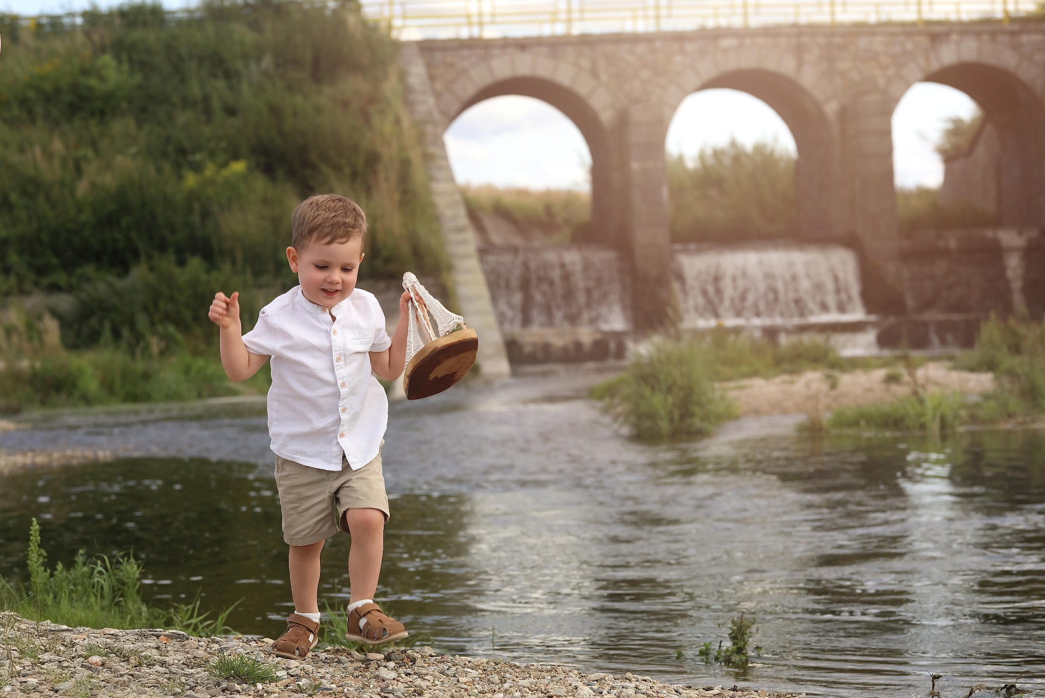 Chłopiec w białej koszuli i beżowych szortach trzyma drewniany model żaglówki na tle rzeki i kamiennego mostu z trzema łukami w letnim, słonecznym dniu.