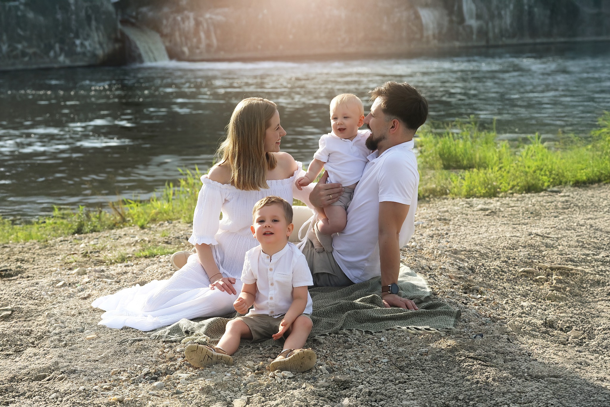 Rodzinna sesja zdjęciowa nad rzeką z wodospadem w tle. Mama w białej sukience, tata trzyma niemowlę, a starszy syn siedzi na kocu. Naturalne światło, spokojna atmosfera.