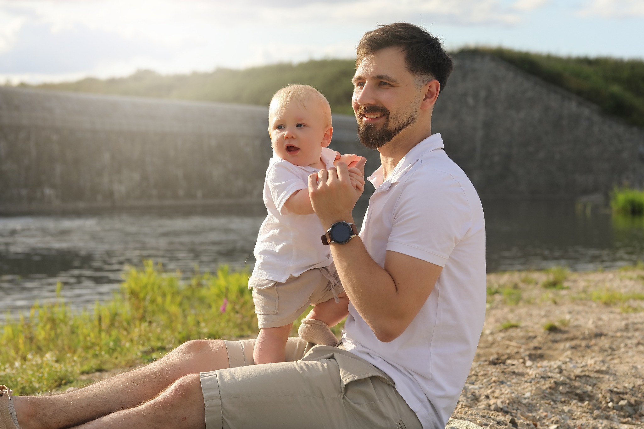Uśmiechnięty tata w beżowych szortach trzymający na kolanach roześmianego synka w plenerze z wodą i zielenią w tle, oboje ubrani na biało.
