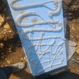 HA-BUD SPÓŁKA Z OGRANICZONĄ ODPOWIEDZIALNOŚCIĄ - Błękitny styropian z klejem w ziemi, widok z góry. Prace izolacyjne fundamentów w Warszawie. But robotnika w lewym dolnym rogu kadru.