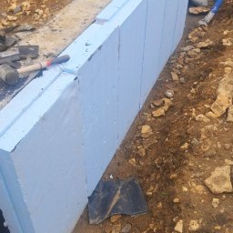 HA-BUD SPÓŁKA Z OGRANICZONĄ ODPOWIEDZIALNOŚCIĄ - Budowa ściany z paneli izolacyjnych z niebieskiego styropianu, widoczne narzędzia budowlane i fragment wykopu. Izolacja termiczna fundamentów.