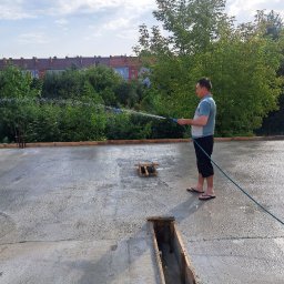 HA-BUD SPÓŁKA Z OGRANICZONĄ ODPOWIEDZIALNOŚCIĄ - Człowiek podlewa świeżo wylaną posadzkę na budowie, używając węża ogrodowego. Widoczne drewniane szalunki i budynki w tle.