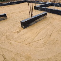 HA-BUD SPÓŁKA Z OGRANICZONĄ ODPOWIEDZIALNOŚCIĄ - Fundamenty domu w trakcie budowy, widoczne zbrojenia i izolacja. Podłoże piaszczyste z wyraźnymi śladami opon. Ujęcie z góry, ukazujące przygotowany teren pod wylewkę.