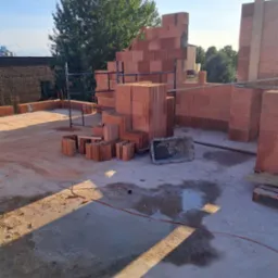 Wznoszenie mur&oacute;w z czerwonej ceramiki na budowie. Widoczne pustaki, rusztowanie i narzędzia. Częściowo wzniesione ściany w słoneczny dzień.