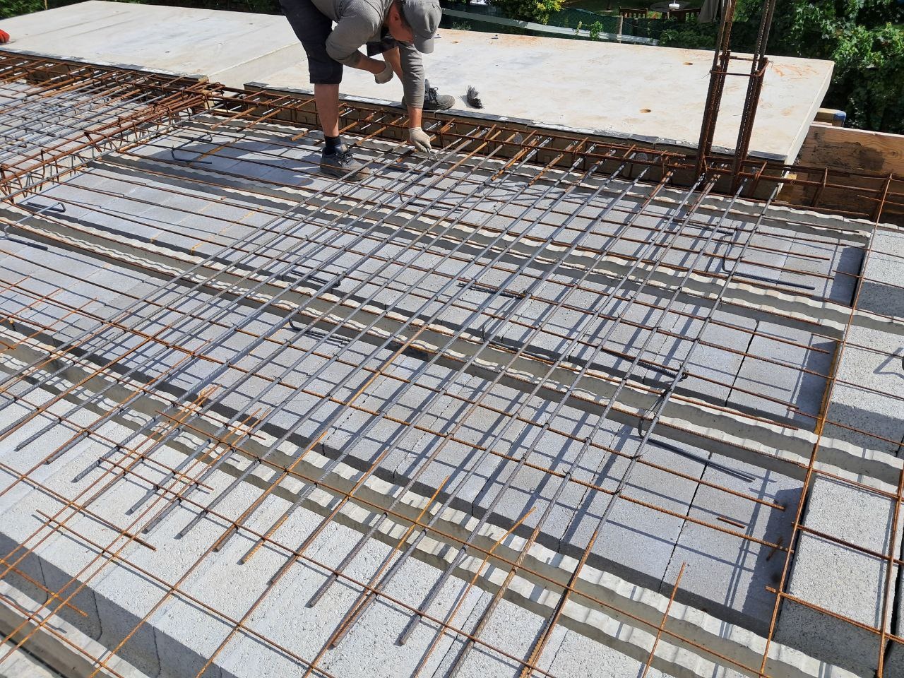 Prace zbrojarskie na budowie: siatka stalowa ułożona na betonowych bloczkach stropowych. Zbrojarz wiąże druty. Widok z góry, w tle zieleń.