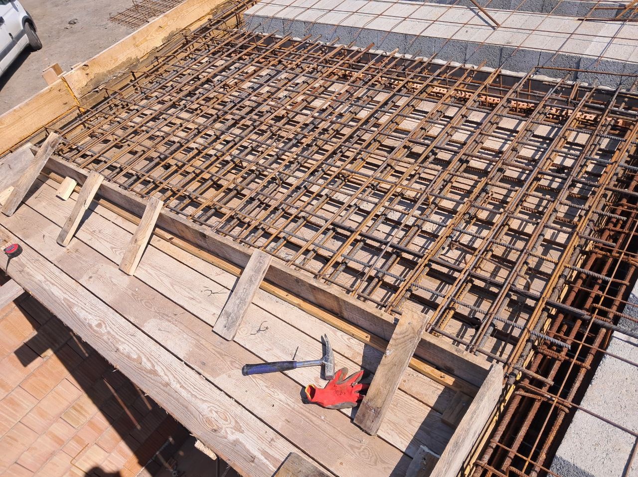 Siatka zbrojeniowa ułożona na drewnianym szalunku, widoczne narzędzia: młotek i rękawice. Przygotowanie do wylewki na budowie.
