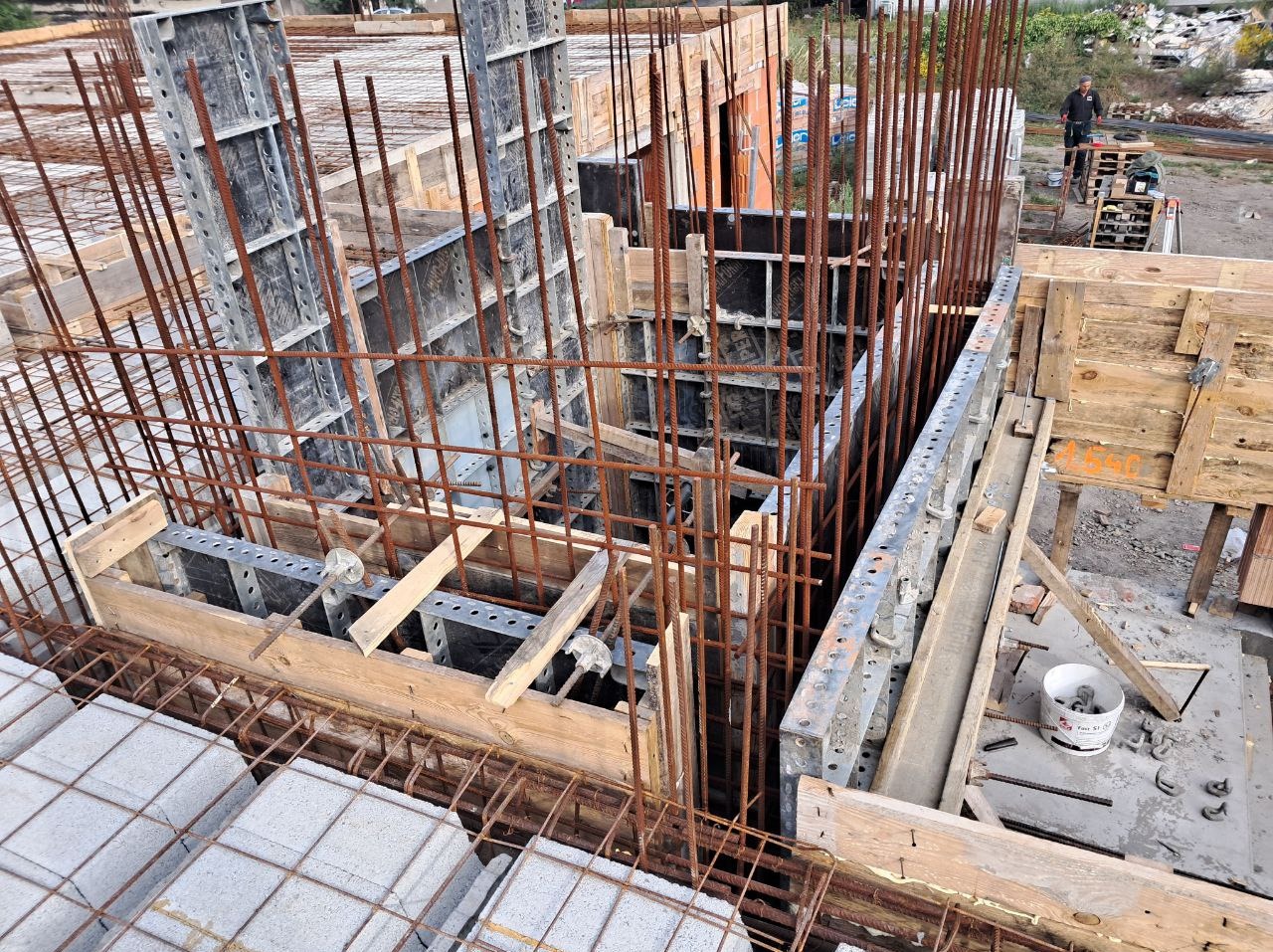 Szare bloki i rustykalne, brązowe pręty zbrojeniowe w szalunkach tworzą konstrukcję budynku w trakcie budowy. Widoczne narzędzia i elementy drewniane.