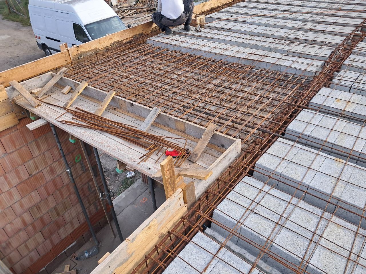 Zbrojenie stropu Teriva z betonowymi pustakami, widok z góry. Na pierwszym planie drewniane szalunki i przygotowane pręty zbrojeniowe. W tle biały samochód dostawczy.