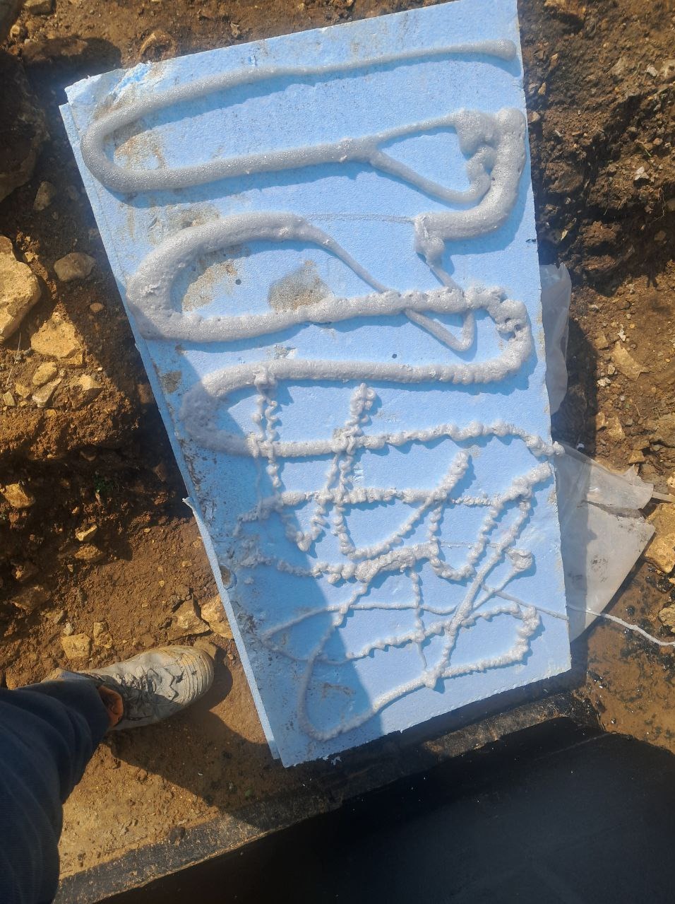 Błękitny styropian z klejem w ziemi, widok z góry. Prace izolacyjne fundamentów w Warszawie. But robotnika w lewym dolnym rogu kadru.