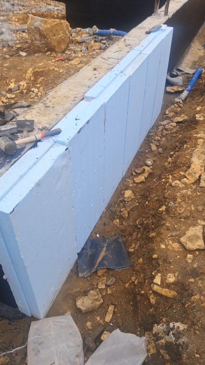 Budowa ściany z paneli izolacyjnych z niebieskiego styropianu, widoczne narzędzia budowlane i fragment wykopu. Izolacja termiczna fundamentów.