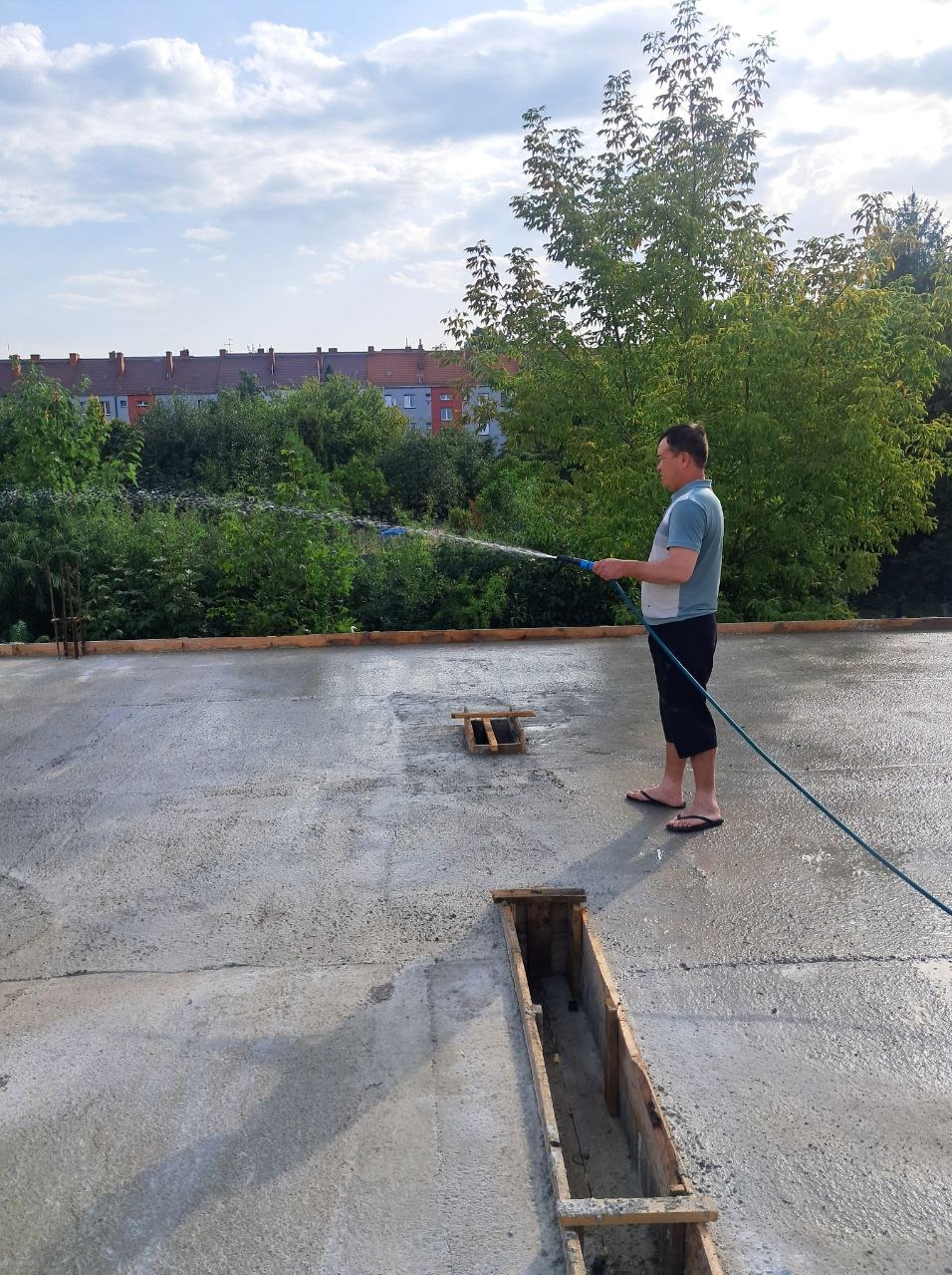 Człowiek podlewa świeżo wylaną posadzkę na budowie, używając węża ogrodowego. Widoczne drewniane szalunki i budynki w tle.