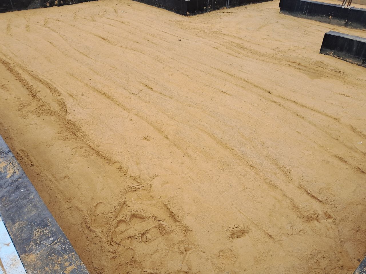 Przygotowany teren pod wylewkę fundamentów z widocznymi śladami stóp i maszyn, otoczony szalunkiem. Piasek wyrównany, gotowy do dalszych prac budowlanych.