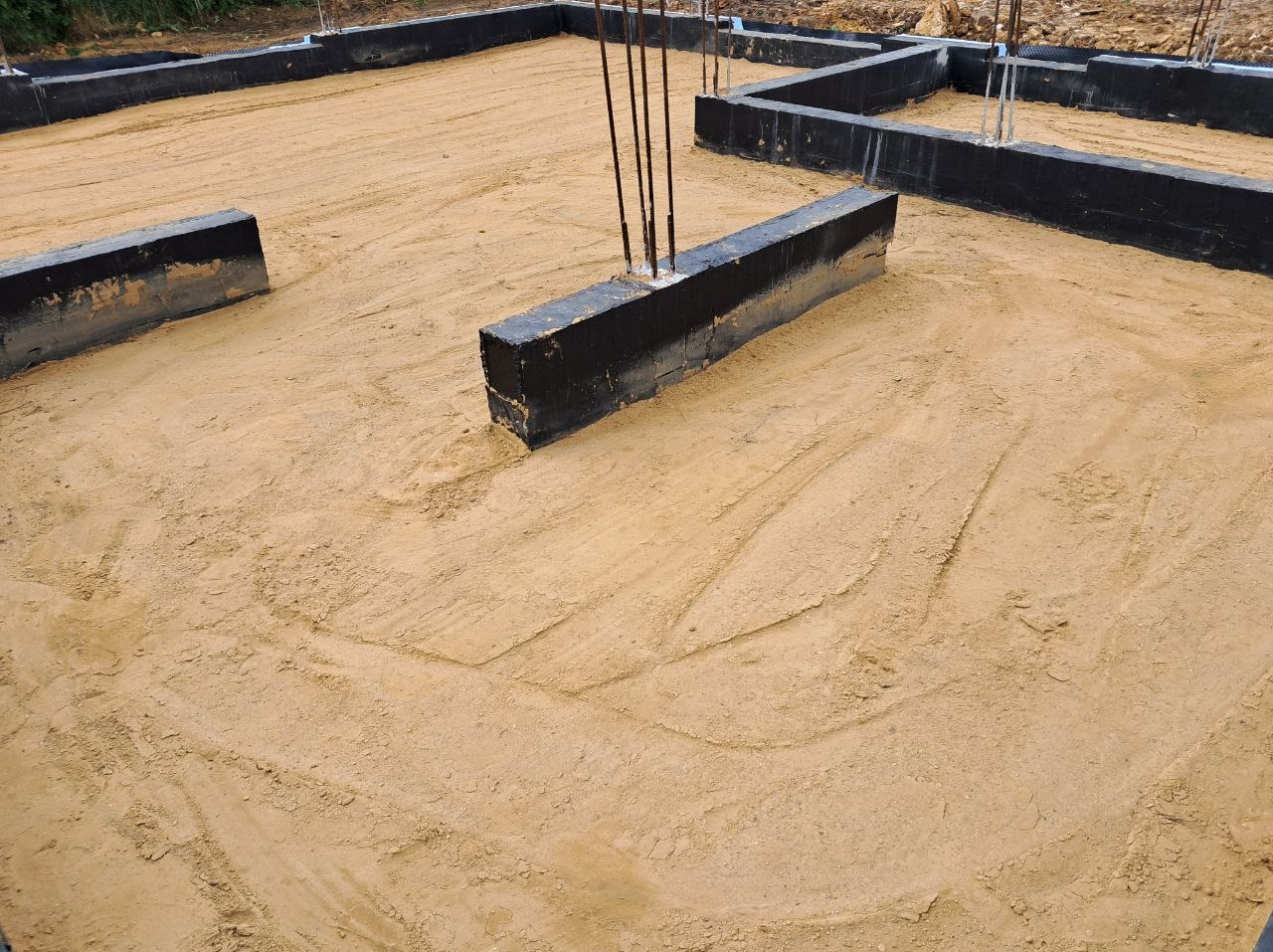 Fundamenty domu w trakcie budowy, widoczne zbrojenia i izolacja. Podłoże piaszczyste z wyraźnymi śladami opon. Ujęcie z góry, ukazujące przygotowany teren pod wylewkę.