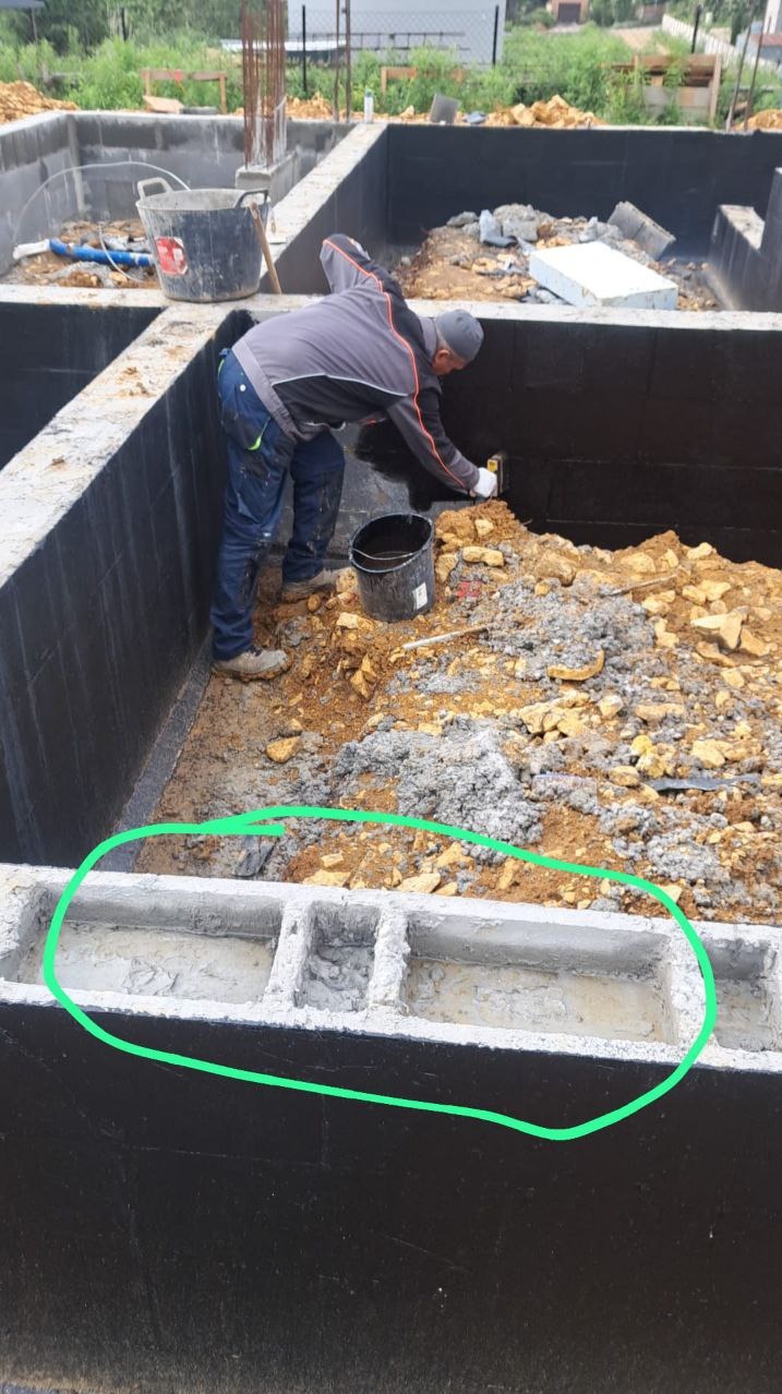 Fundamenty budynku w trakcie izolacji bitumicznej, widoczne bloczki betonowe i pracownik zabezpieczający ściany. Ujęcie z góry, naturalne światło.