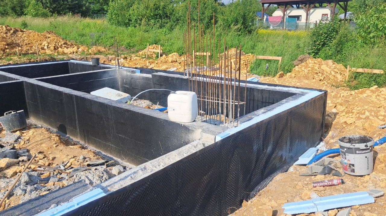 Fundamenty budynku z izolacją przeciwwilgociową i wystającymi prętami zbrojeniowymi, widoczne materiały budowlane i teren budowy.