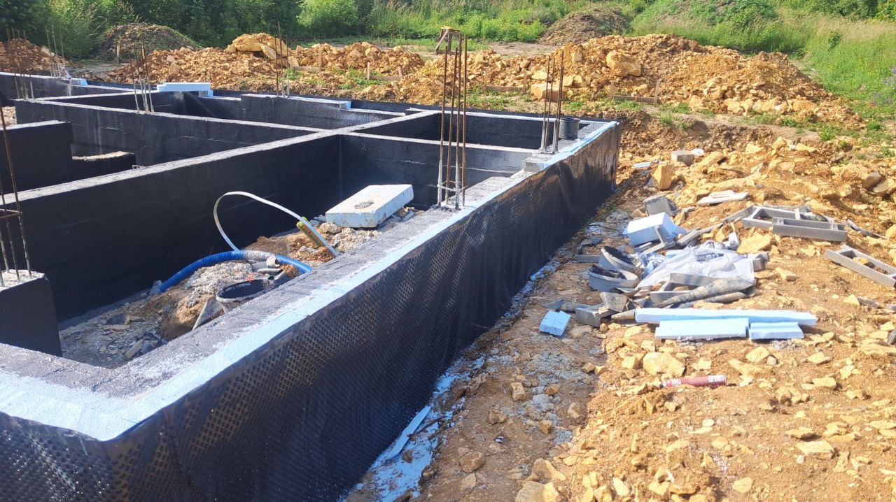 Zabezpieczone fundamenty budynku z wystającymi prętami zbrojeniowymi, widoczne materiały budowlane i ziemia w tle. Izolacja przeciwwilgociowa.