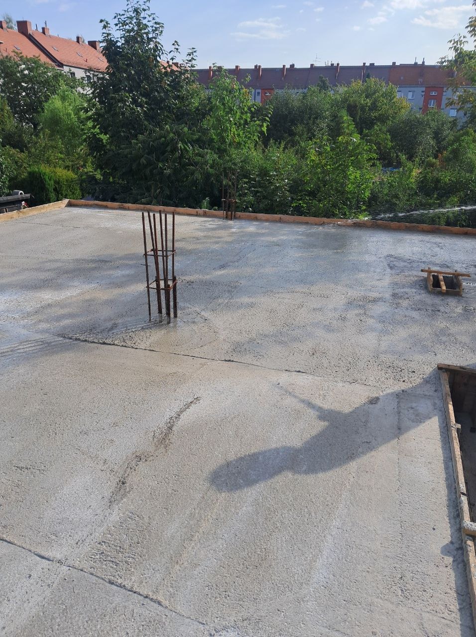 Świeżo wylana, gładka wylewka betonowa z wystającymi prętami zbrojeniowymi, otoczona drewnianym szalunkiem. W tle zieleń drzew i budynki mieszkalne.