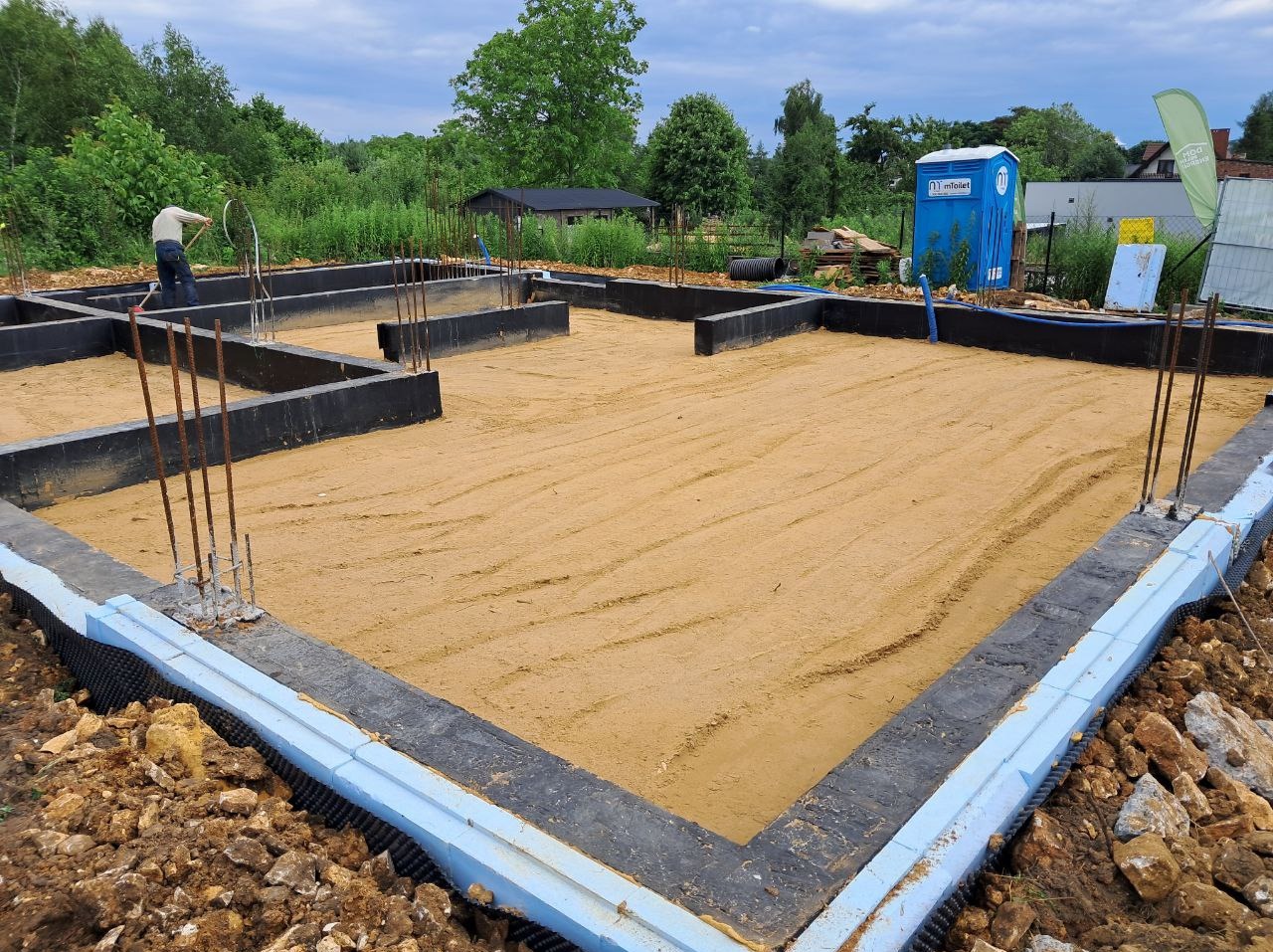 Przygotowany fundament z izolacją i wystającymi prętami zbrojeniowymi, piasek zasypany wewnątrz, toaleta TOI-TOI w tle, widok z bliska.