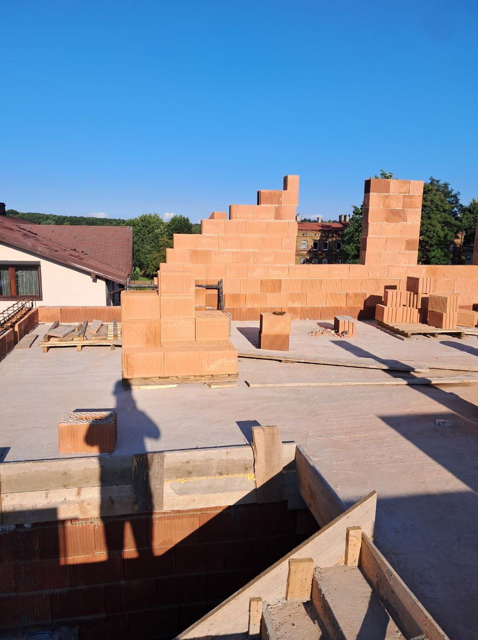 Wznoszenie murów z czerwonej cegły ceramicznej na betonowej płycie stropowej. Widoczny cień osoby, dom w tle i błękitne niebo.