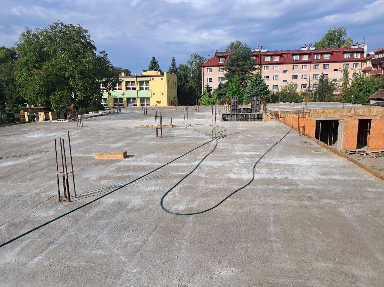 Wylana płyta fundamentowa z wystającymi prętami zbrojeniowymi i częściowo wymurowanymi ścianami. W tle budynki mieszkalne i drzewa, słoneczny dzień.