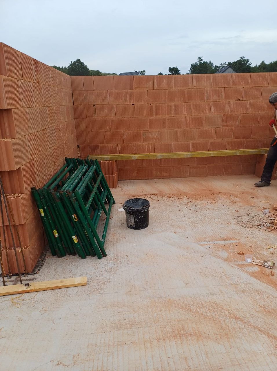 Wznoszenie ściany z czerwonych bloczków na budowie. Widoczne rusztowania, poziomica i częściowo postać robotnika. Ściana w trakcie budowy, z widoczną fakturą bloczków.