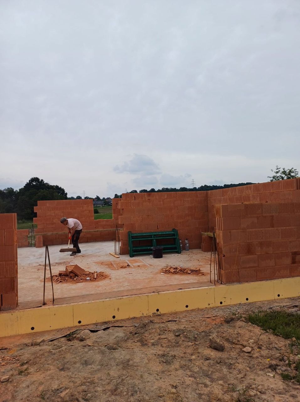 Wnętrze powstającego domu z czerwonej cegły. Murarz w pracy, widoczne ściany i podłoże. W tle zieleń i pochmurne niebo.