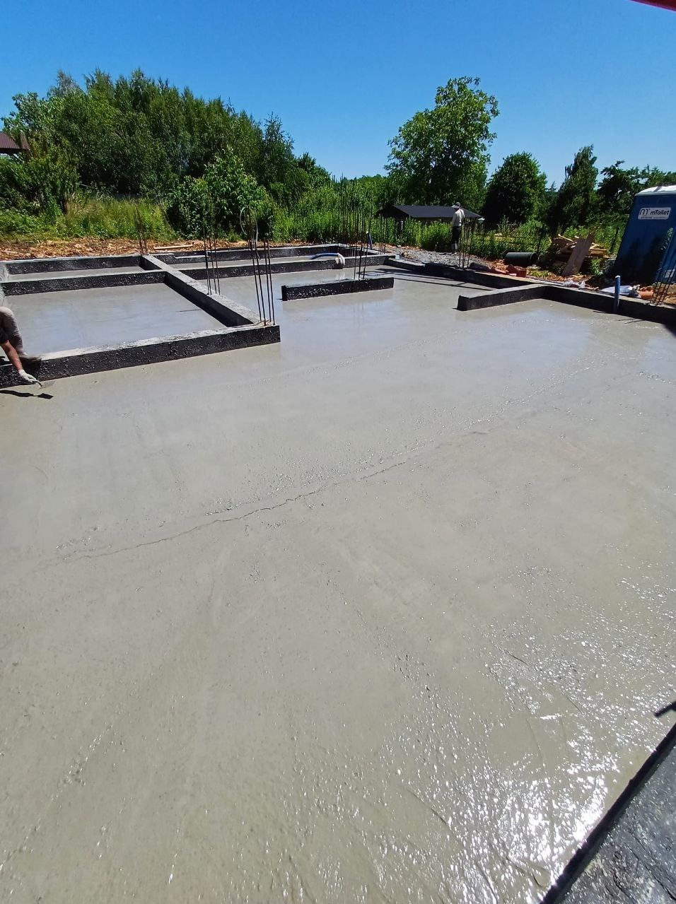 Świeża, jasna wylewka fundamentowa na budowie w słoneczny dzień. Widoczne wystające pręty zbrojeniowe i częściowo pracownik. W tle zieleń drzew.