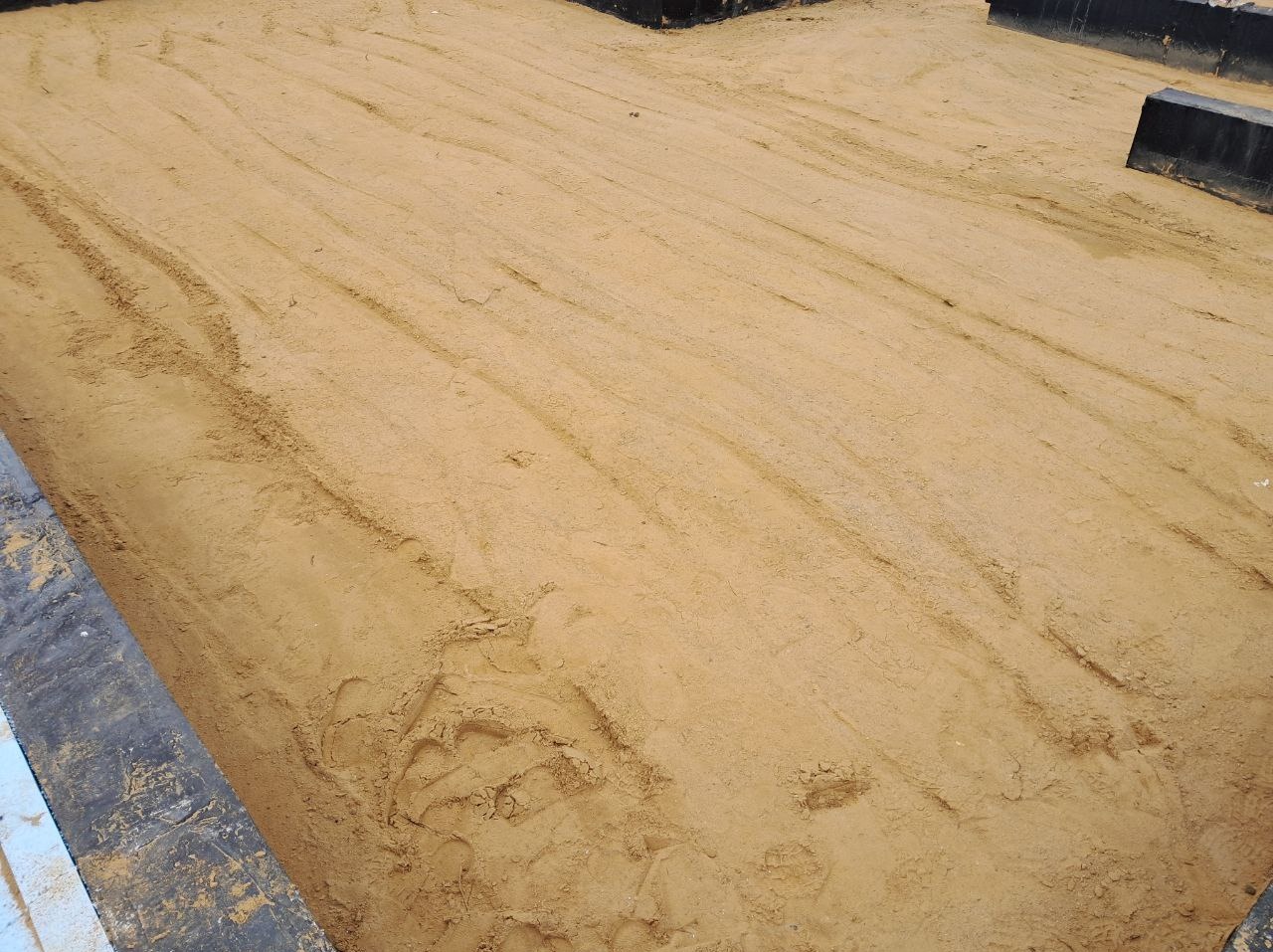 Przygotowany piasek pod wylewkę, otoczony czarną formą, widoczne ślady stóp i opon, gotowy do dalszych prac budowlanych.