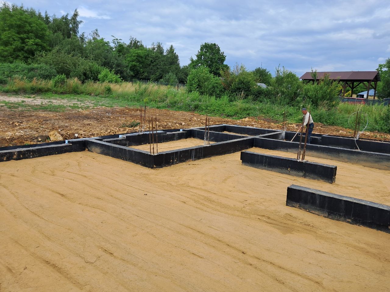 Fundamenty domu jednorodzinnego z wystającymi prętami zbrojeniowymi na piaszczystym podłożu w słoneczny dzień. W tle widoczna zieleń i drewniana altana.