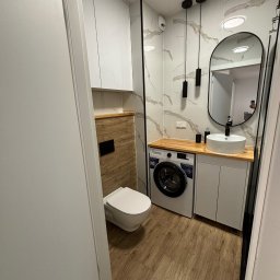 Firma Usługowa BUILD&GO Tomasz Wydra - Nowoczesna łazienka z marmurowymi płytkami, drewnianym blatem, owalnym lustrem i pralką pod umywalką. Minimalistyczne oświetlenie i podłoga imitująca drewno.