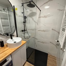 Firma Usługowa BUILD&GO Tomasz Wydra - Nowoczesna łazienka z kabiną typu walk-in, białą umywalką na drewnianym blacie i grzejnikiem drabinkowym. Marmurowe płytki na ścianach i czarny brodzik.