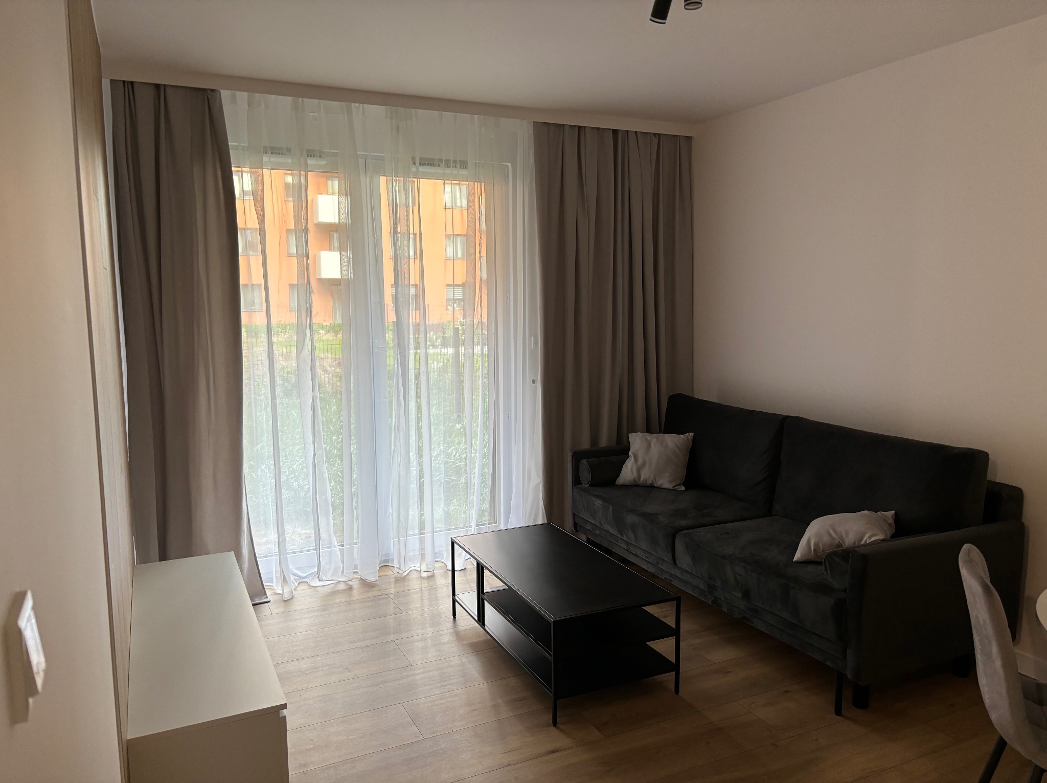 Nowoczesny salon z szarą sofą, czarnym stolikiem kawowym i jasnymi zasłonami. Widok na budynek z okna. Minimalistyczny design wnętrza.